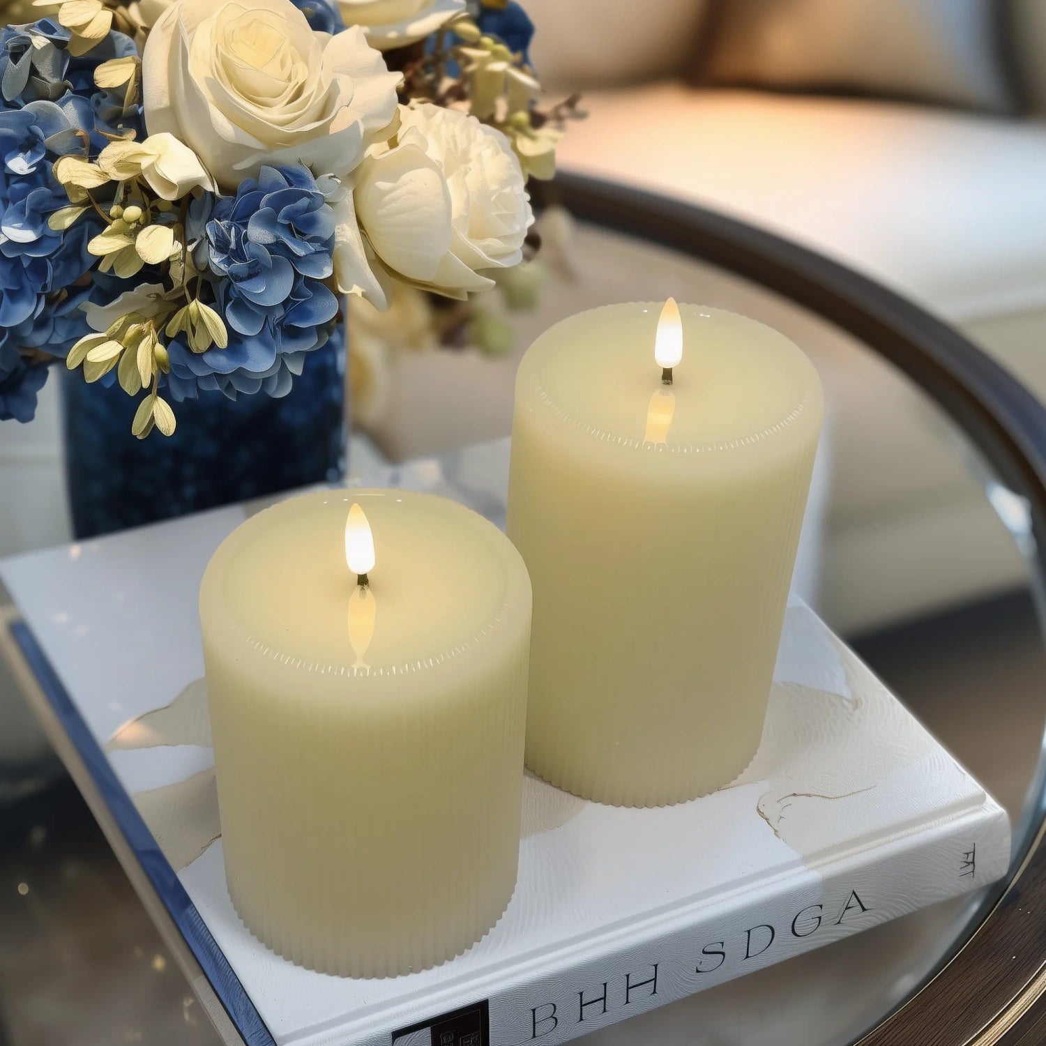 Two lit candles on a book with a decorative flower arrangement in the background.#color_ivory