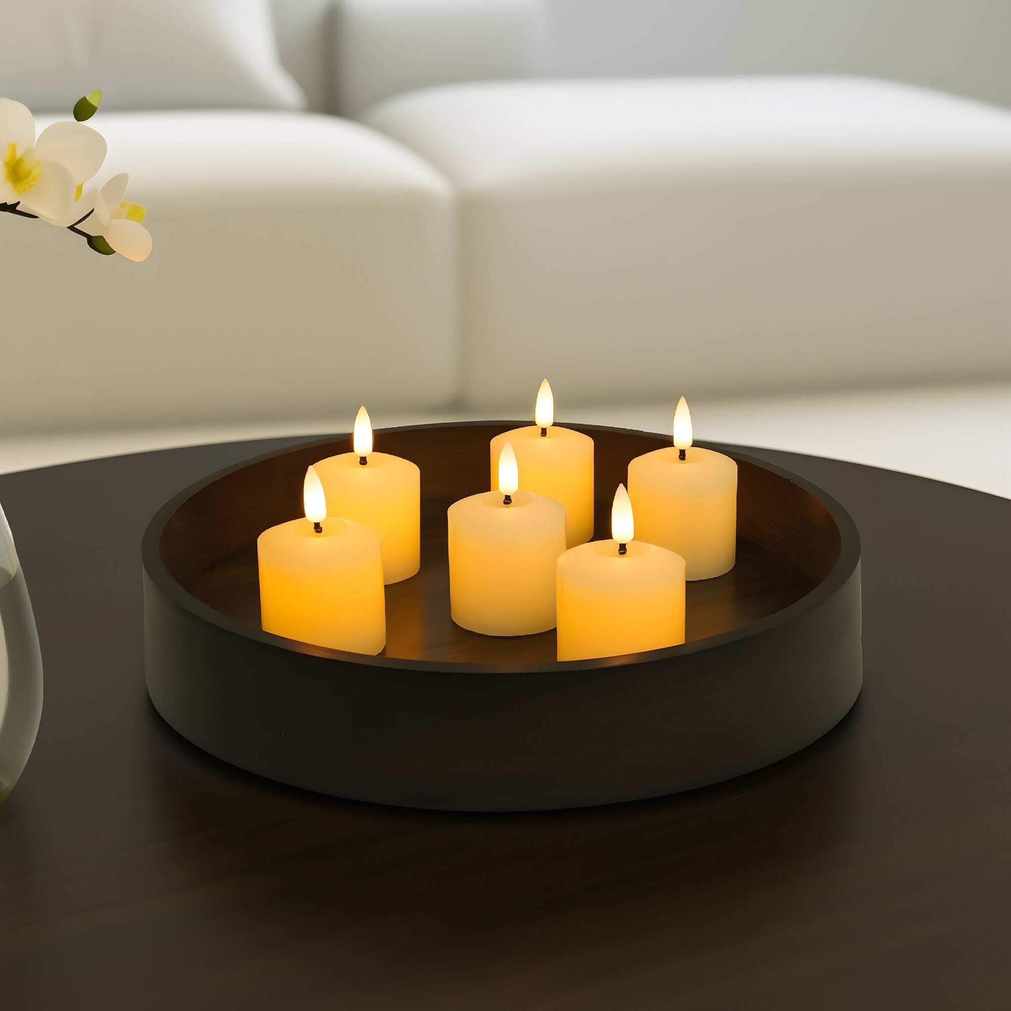 Round black candle holder with six lit candles on a dark surface, with a blurred white couch in the background.