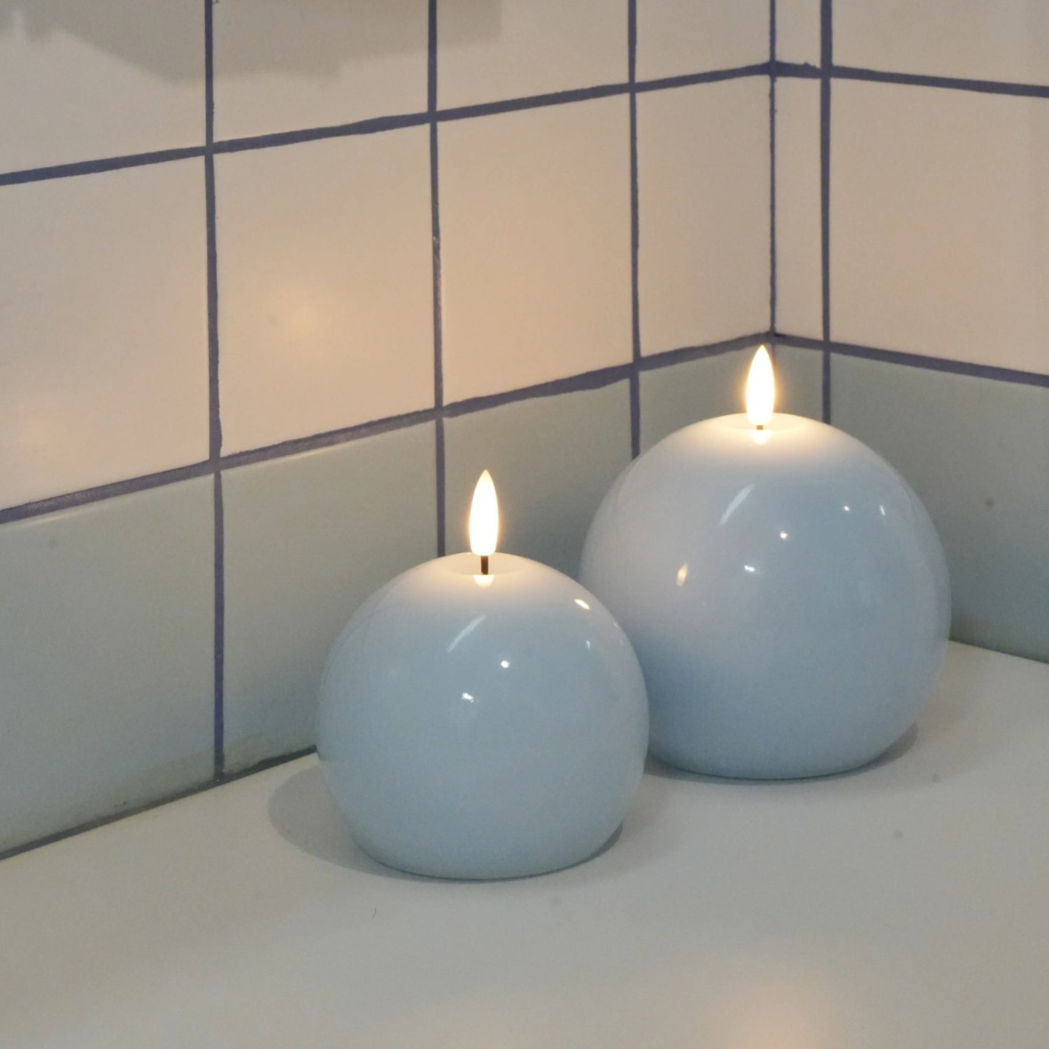 Two spherical candles with flames on a tiled bathroom counter.#color_shiny-blue