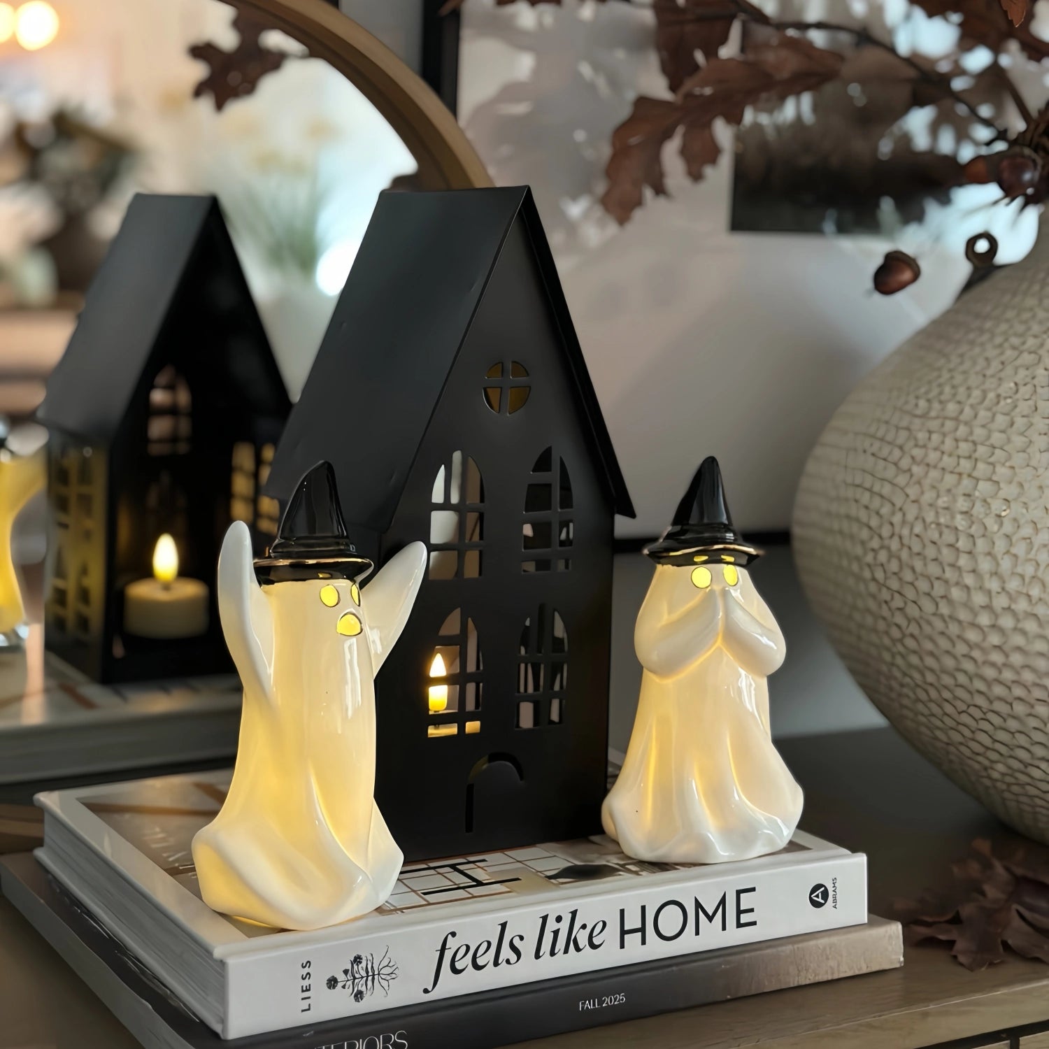 A Halloween-themed tabletop display featuring a black haunted house lantern and two glowing cream ghost figurines with pointed hats, arranged on stacked books in a home decor setting.