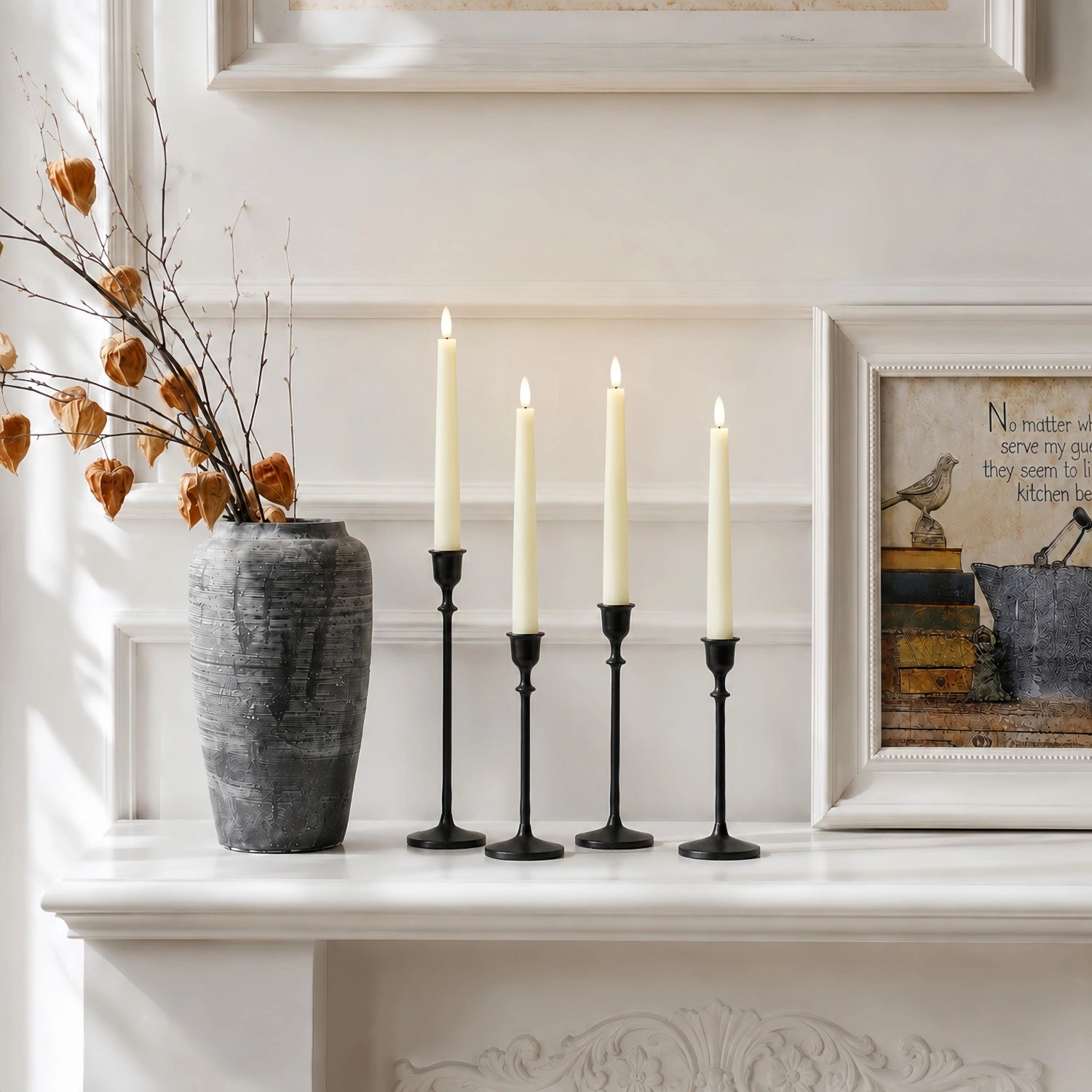 Four black candlestick holders with cream taper candles displayed on a white mantelpiece, styled with a gray textured vase and dried branches.