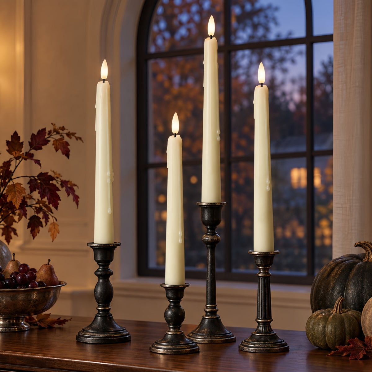 Four cream taper candles in black metal candlesticks arranged on a wooden table in a warmly lit room, with autumn leaves, a bowl of fruit, and pumpkins nearby and a window in the background.