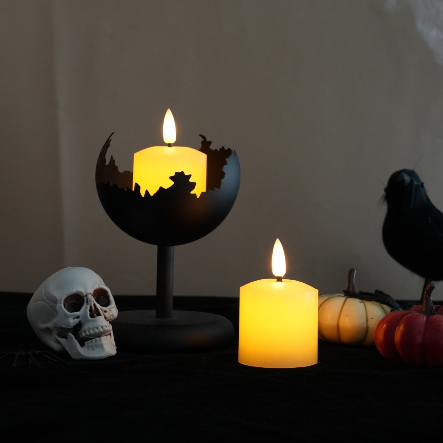 Two lit LED pillar candles displayed on a dark tabletop with Halloween decor, including a skull, small pumpkins, and a black raven silhouette. One candle is in a black globe-style holder with a cutout skyline design, and the other is in a simple black holder.