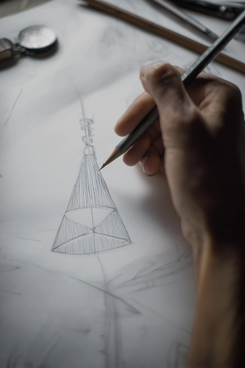 Hand holding a pencil drawing a geometric shape on paper with tools in the background