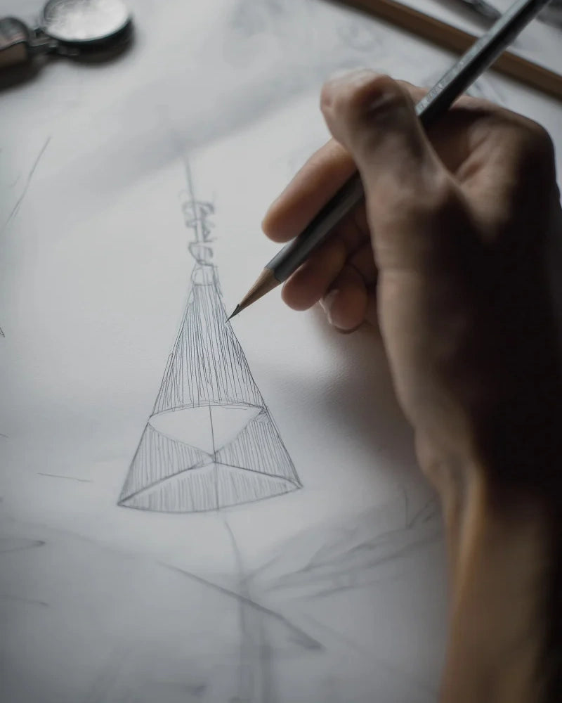 Hand holding a pencil drawing a geometric shape on paper with a spoon in the background.