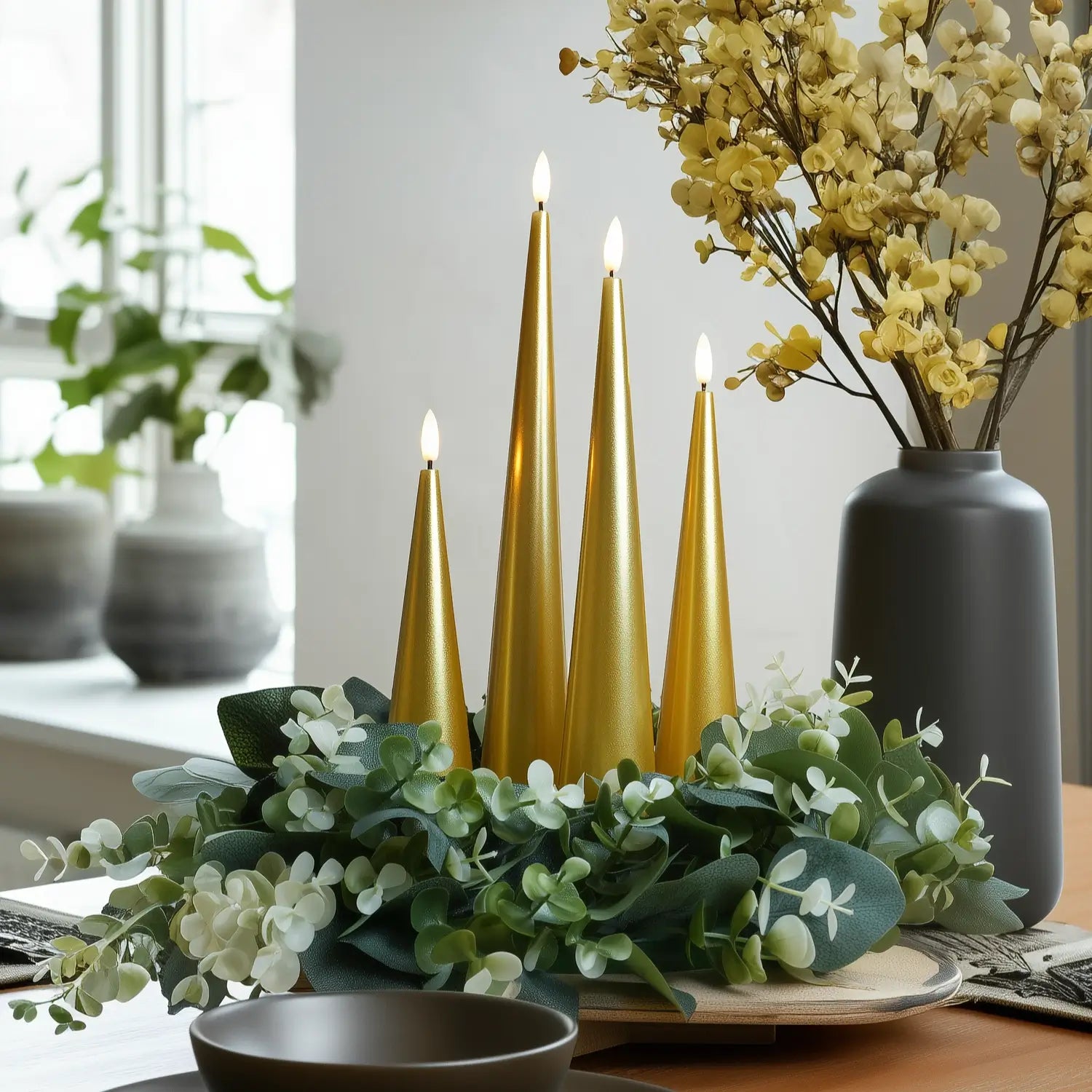 Decorative setup with gold candles, greenery, and a vase with yellow flowers on a table.