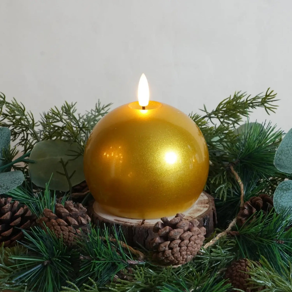 Gold spherical candle on a wooden base surrounded by greenery and pine cones.