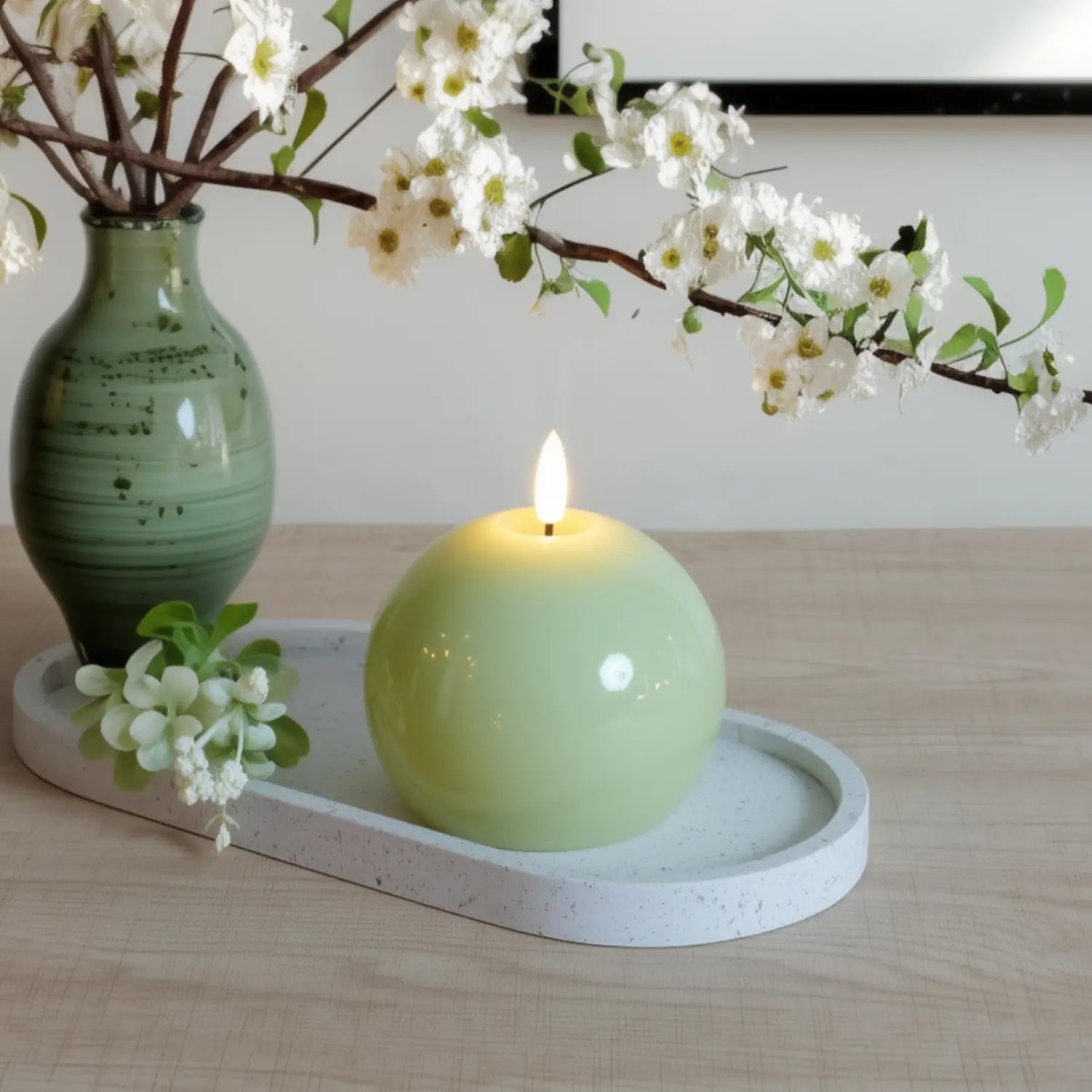 Green candle on a white tray with a vase and floral branches on a wooden surface