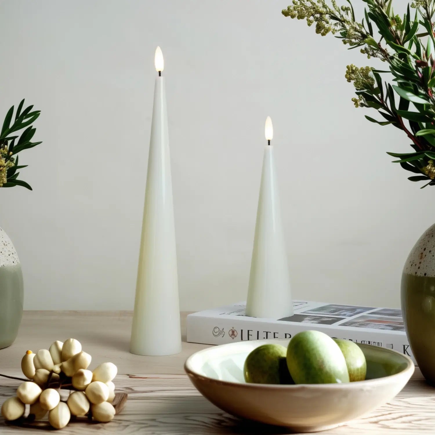 Two white cone-shaped candles on a wooden surface with a bowl of green apples and decorative items.