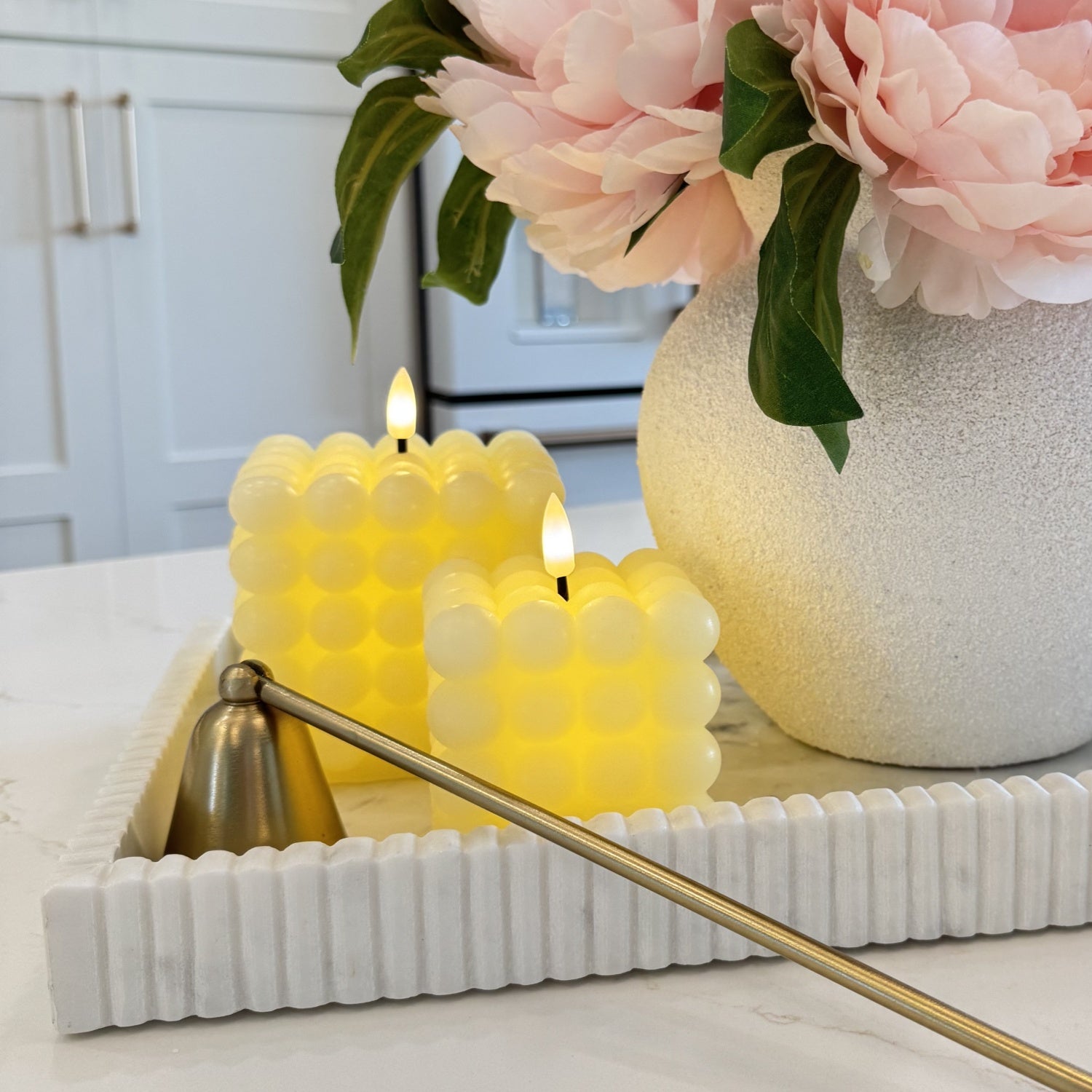 Two lit candles in a decorative holder with a vase of pink flowers in the background.