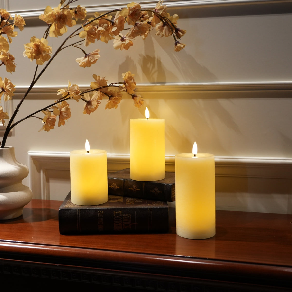 Three lit cream-colored pillar candles arranged on stacked dark books on a wooden table, with decorative dried flowers in a vase against a paneled wall.
