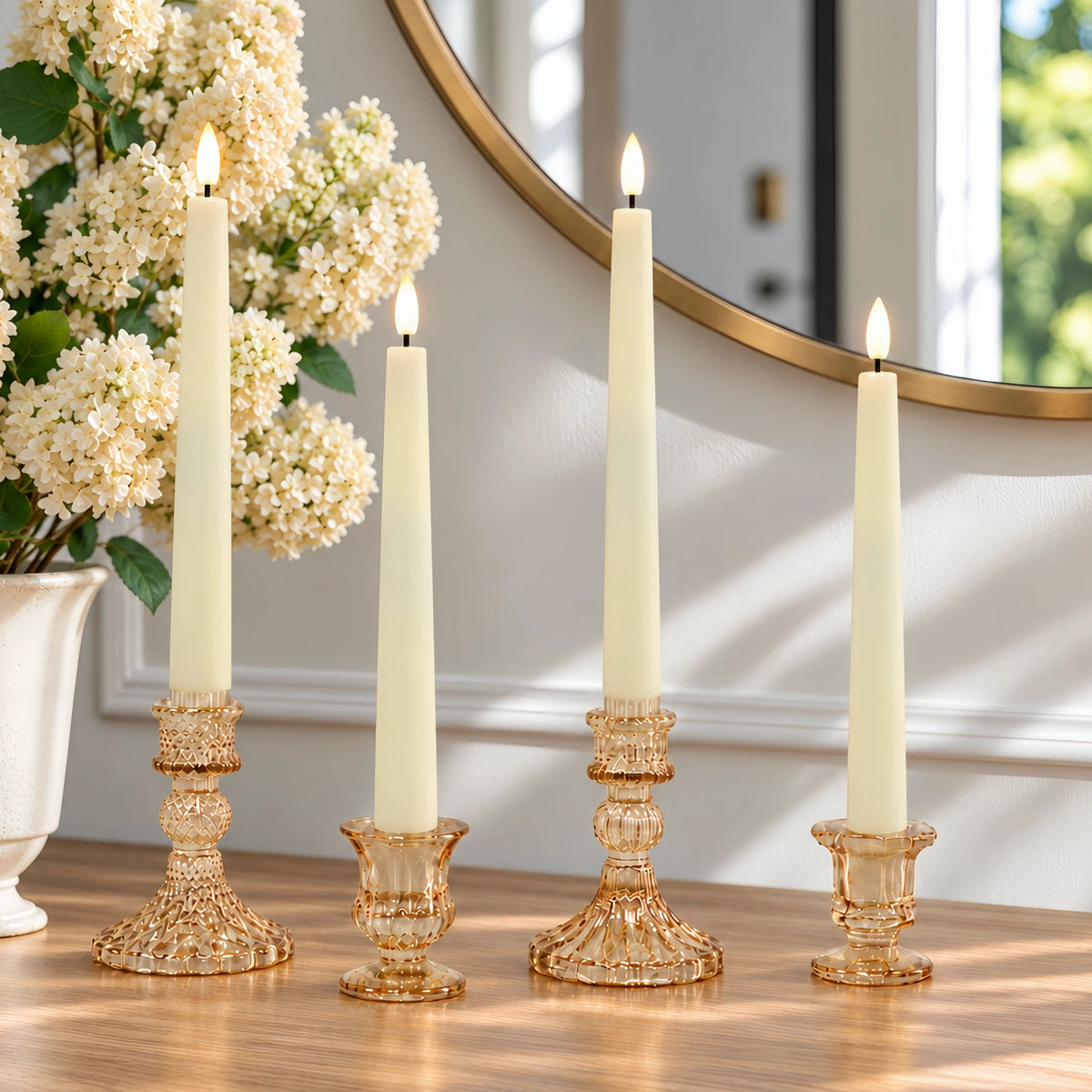 Four lit taper candles in ornate clear glass holders arranged on a wooden table in a bright room, with a vase of white flowers and a round mirror in the background.