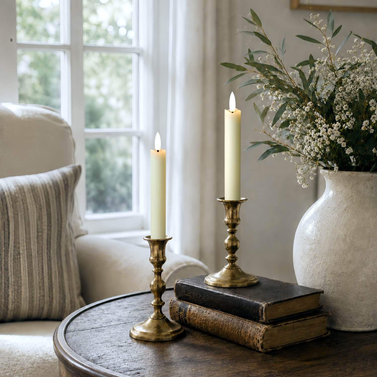 Two lit cream pillar candles in ornate brass candlesticks placed on stacked vintage books on a round wooden table, with a beige armchair and a white vase of white flowers in the softly lit background.#color_rustic-ivory