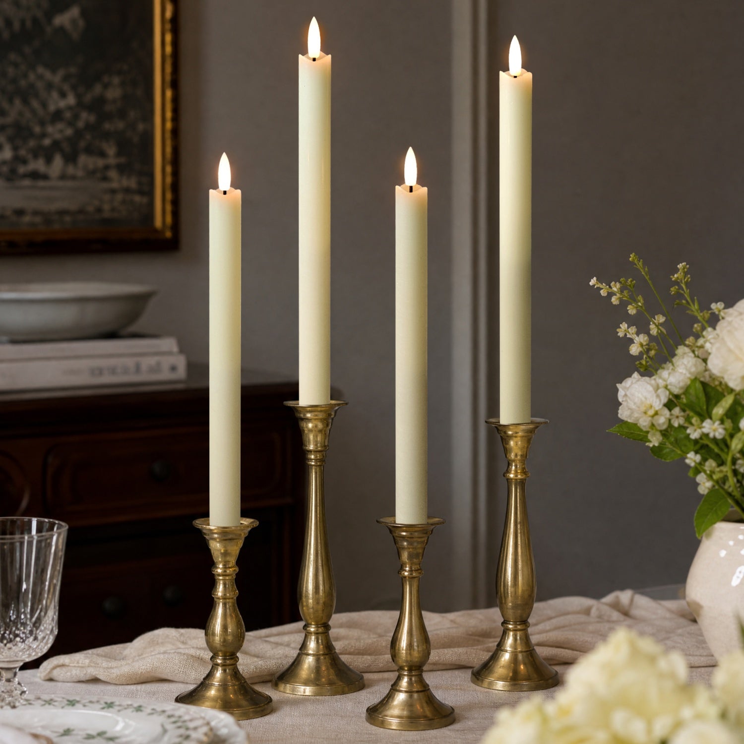 Three lit cream taper candles in ornate gold candlesticks arranged on a table in a styled interior setting.