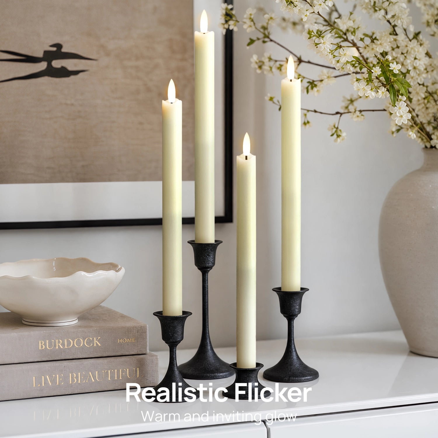 Three black metal candlesticks holding cream pillar candles with realistic flickering flames, styled on a white surface with books, a bowl, framed wall art, and a vase of white blossoms in a bright interior setting.