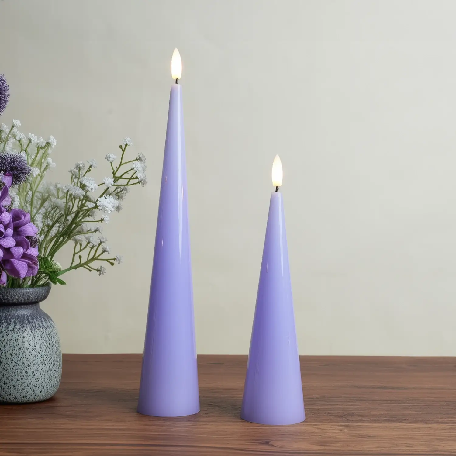 Two purple candles on a wooden surface with a vase of flowers in the background.#color_shiny-purple