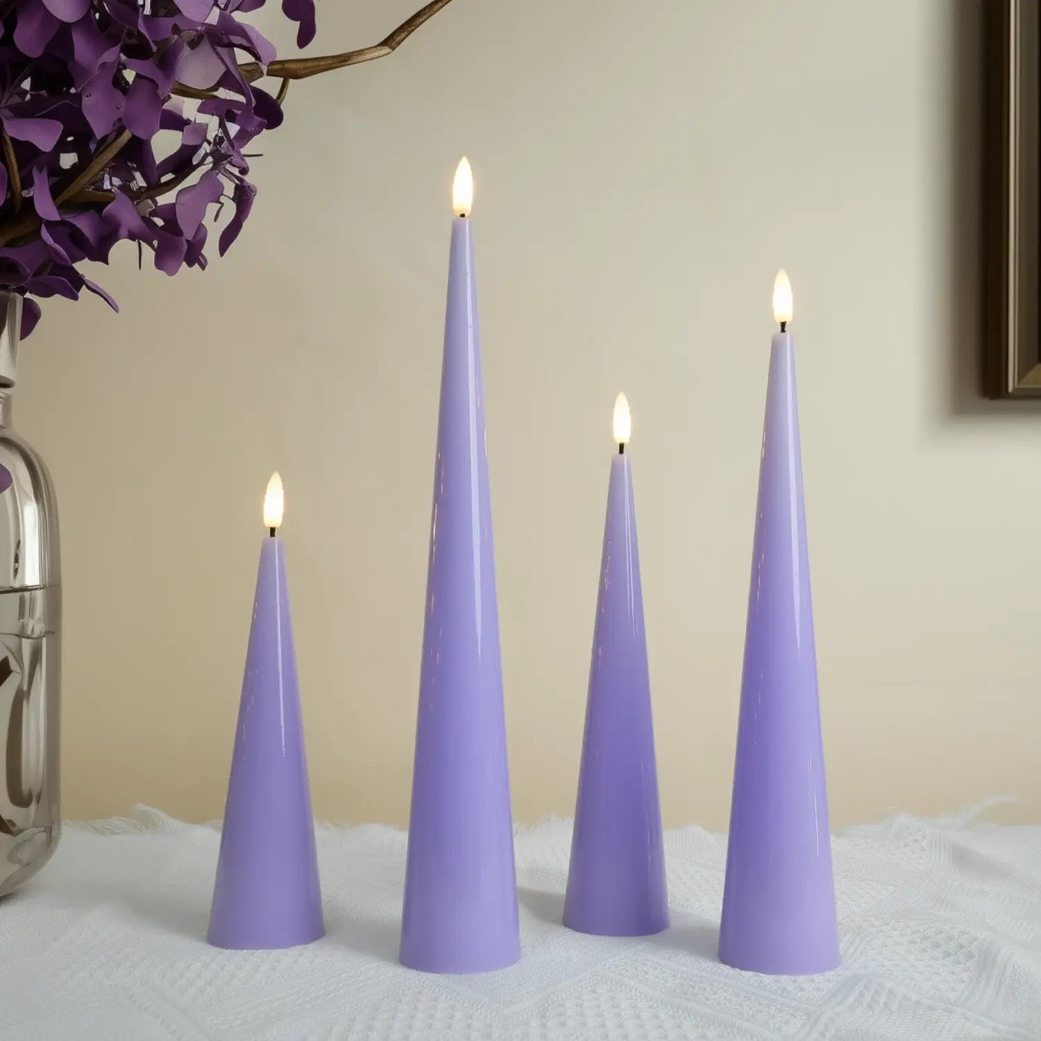 Four purple cone-shaped candles with lit wicks on a textured surface, with a vase of purple flowers in the background.#color_shiny-purple