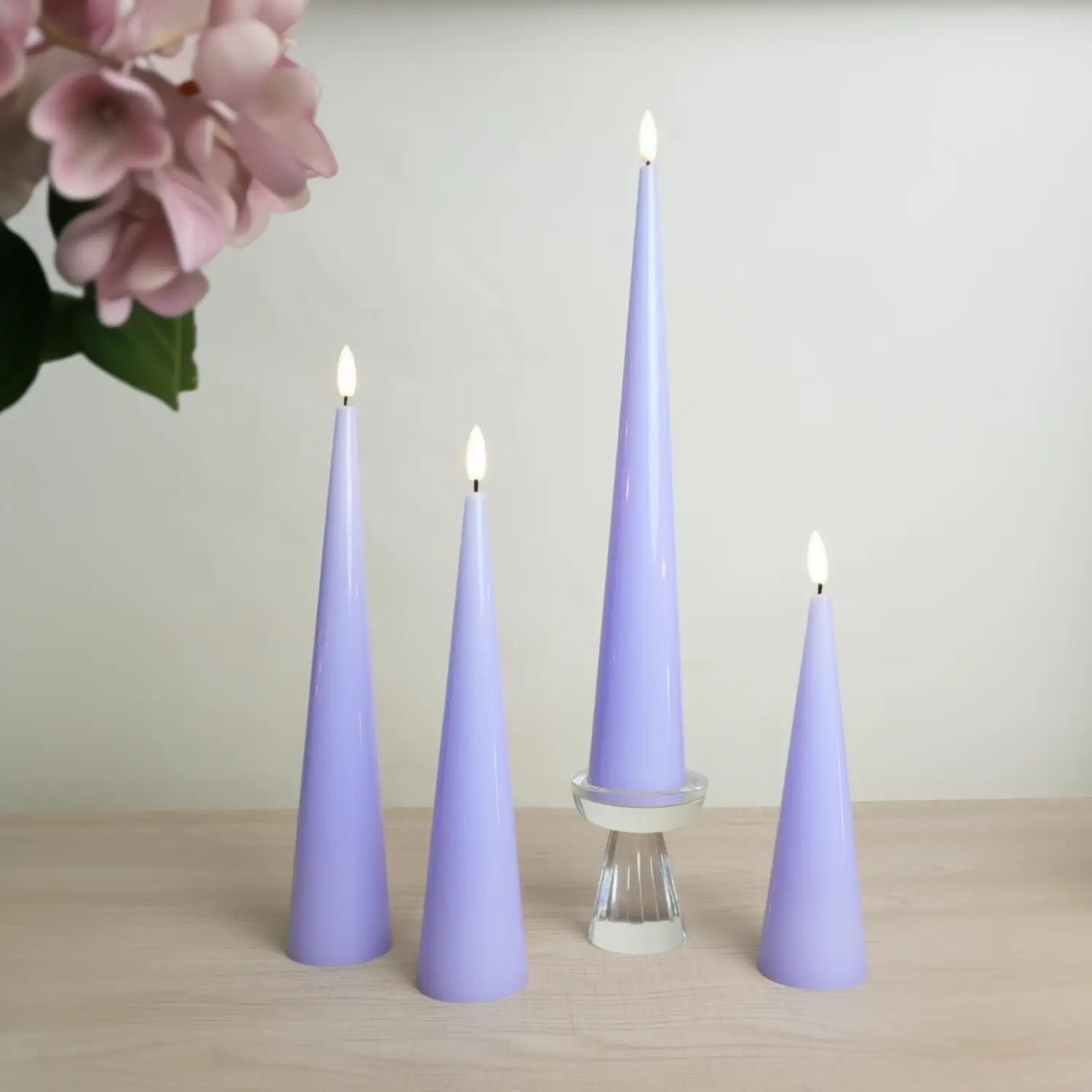 Set of purple cone-shaped candles on a wooden surface with pink flowers in the background.#color_shiny-purple
