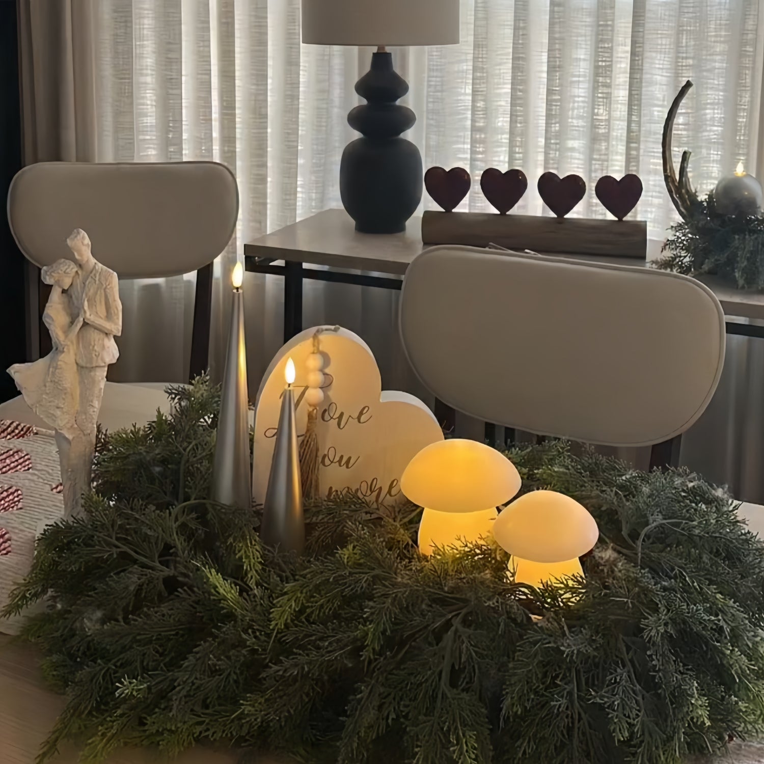 Two lit silver candles on a white surface with a vase and decorative items in the background.
