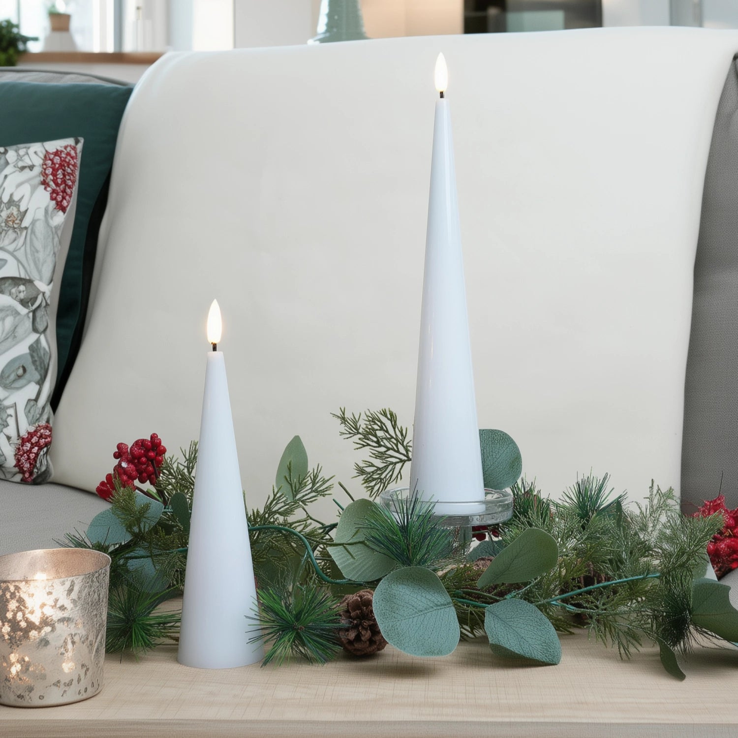 Two white candles on a table with greenery and a decorative pillow in the background#color_shiny-white