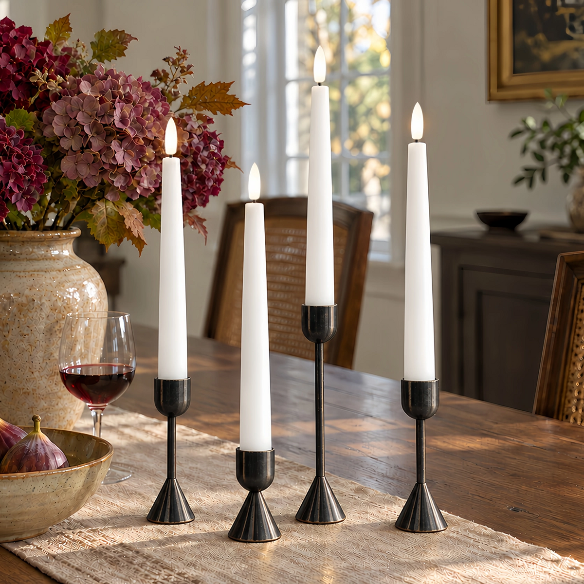 Four black taper candle holders with lit white taper candles arranged on a wooden dining table in a home interior setting, with a vase of flowers, wine glass, and cabinetry in the background.#color_white