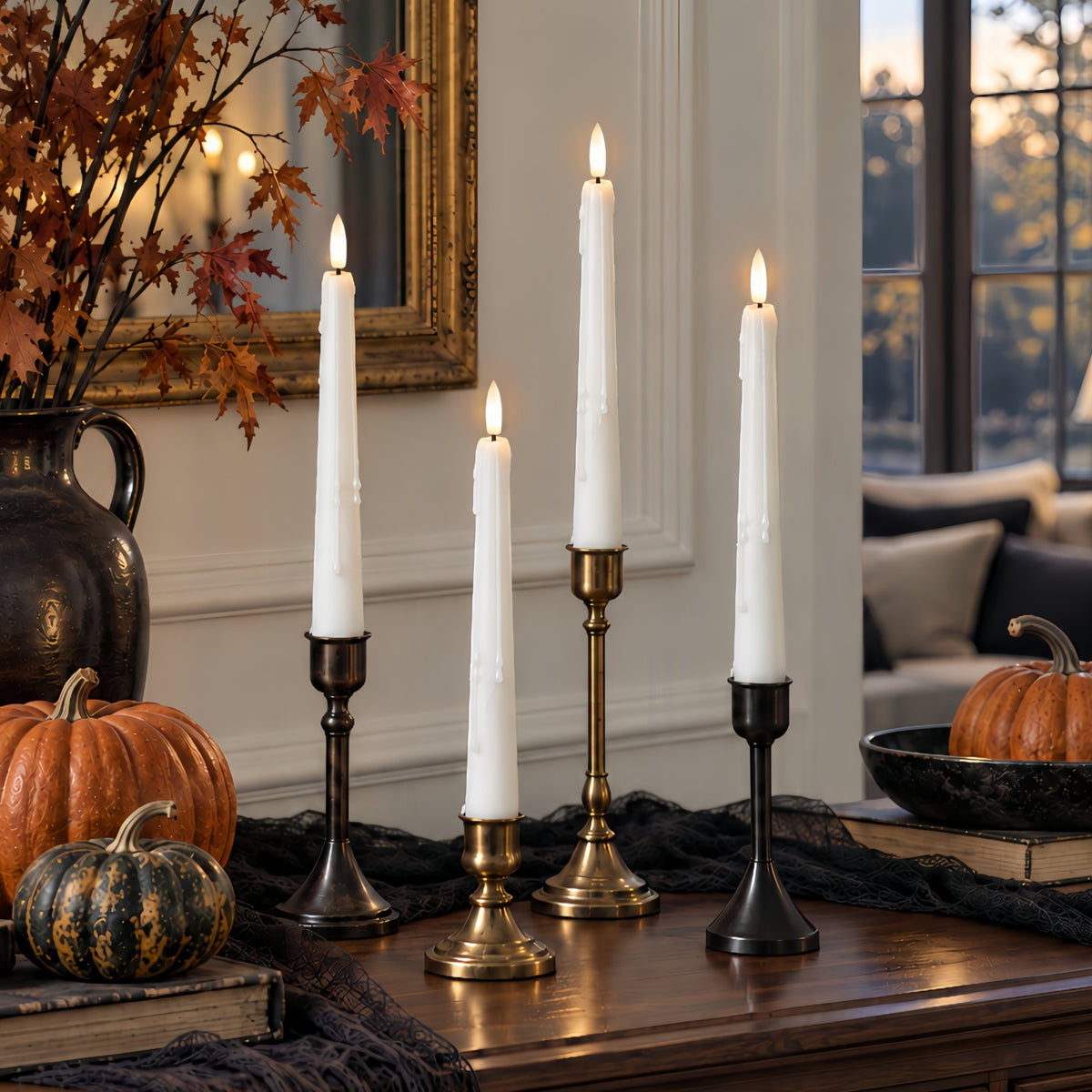 Four lit taper candles in black and brass candlesticks arranged on a wooden table in a styled home setting with pumpkins, a mirror, and a window.
