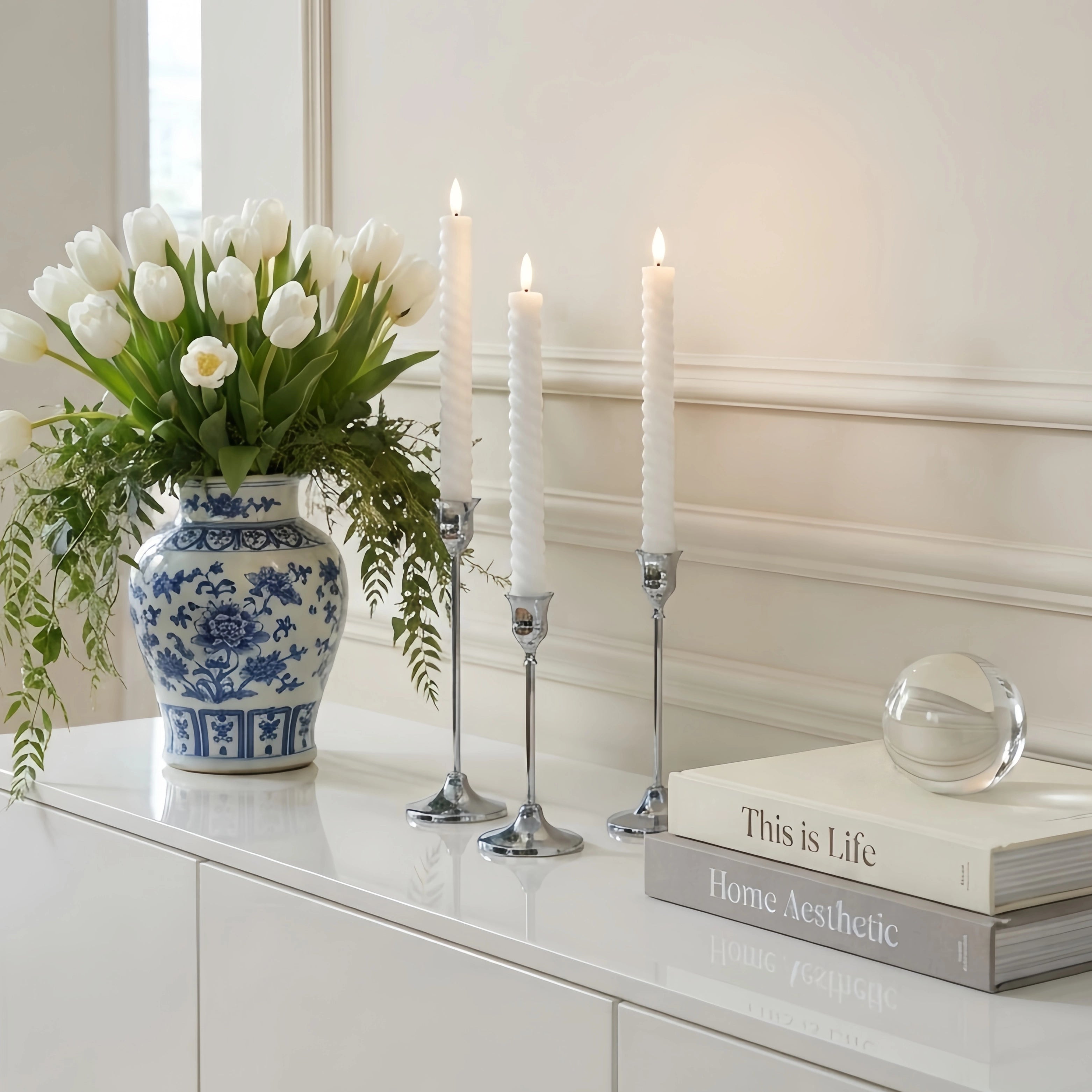 A styled interior scene showing a white ceramic vase filled with white tulips and green foliage on a glossy white cabinet, alongside three lit white taper candles in silver holders, stacked books, and a clear glass orb against a cream paneled wall.