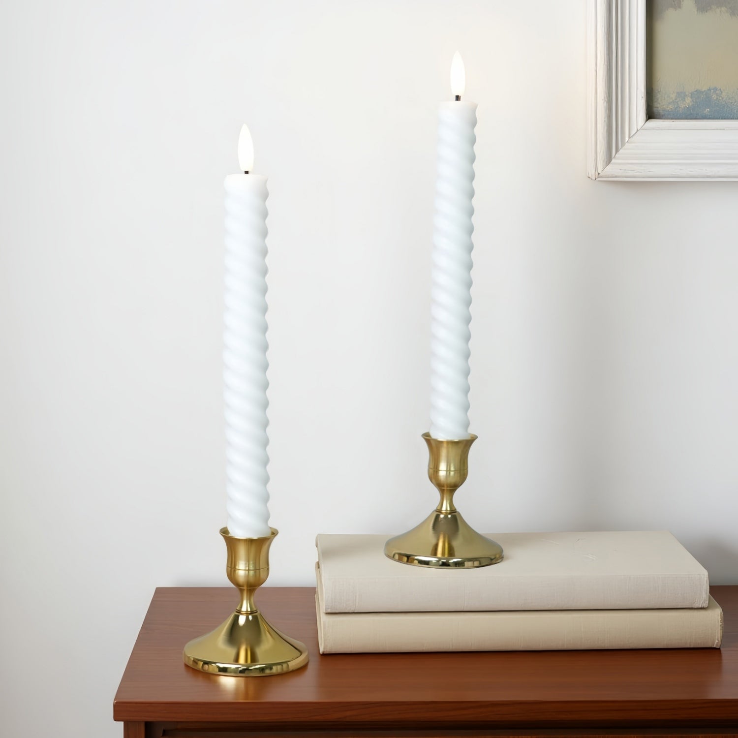 Two white spiral taper candles in gold candlesticks, one standing alone and the other resting on top of two neutral-colored books on a wooden table against a white wall.