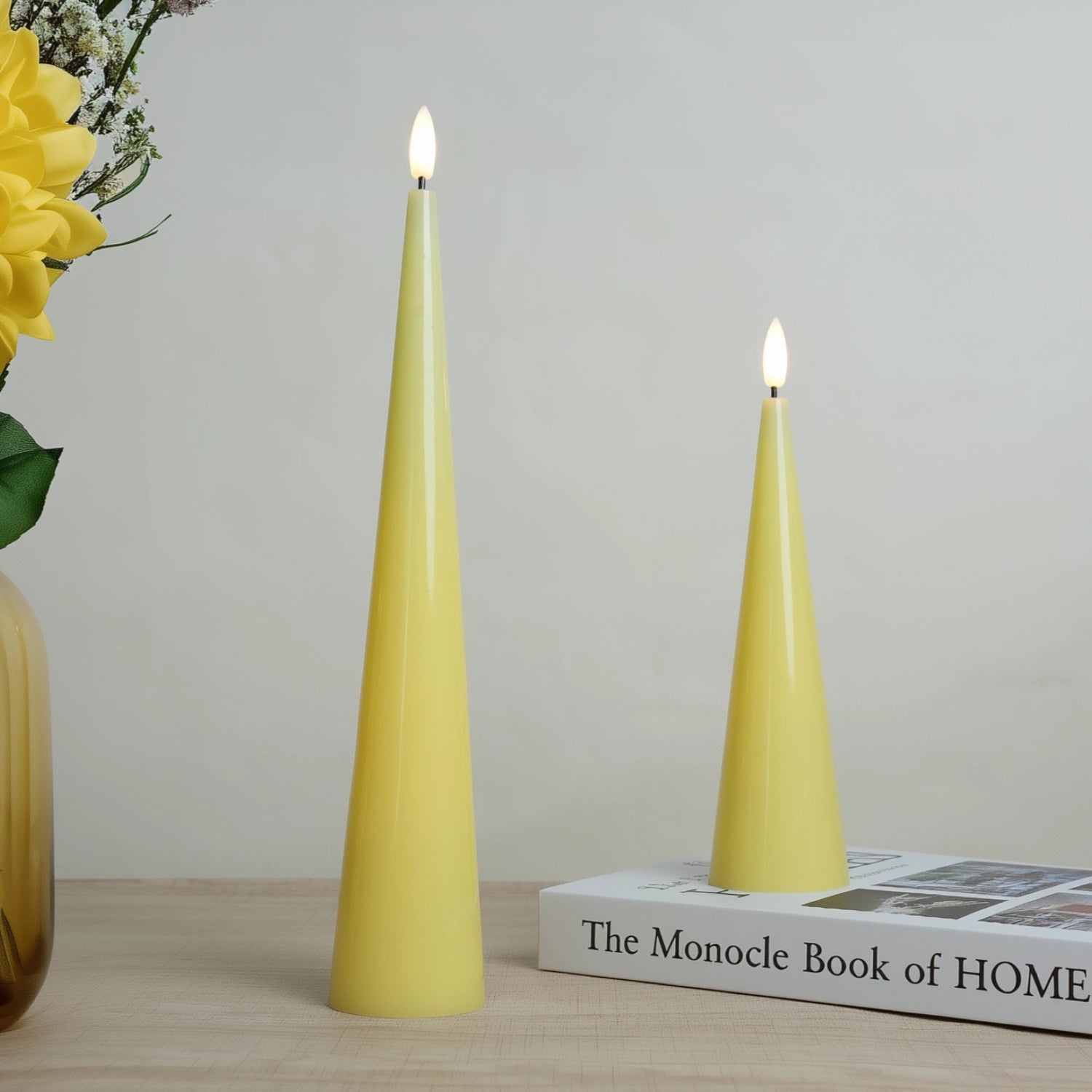 Two yellow candles on a book with a vase of flowers in the background#color_shiny-yellow