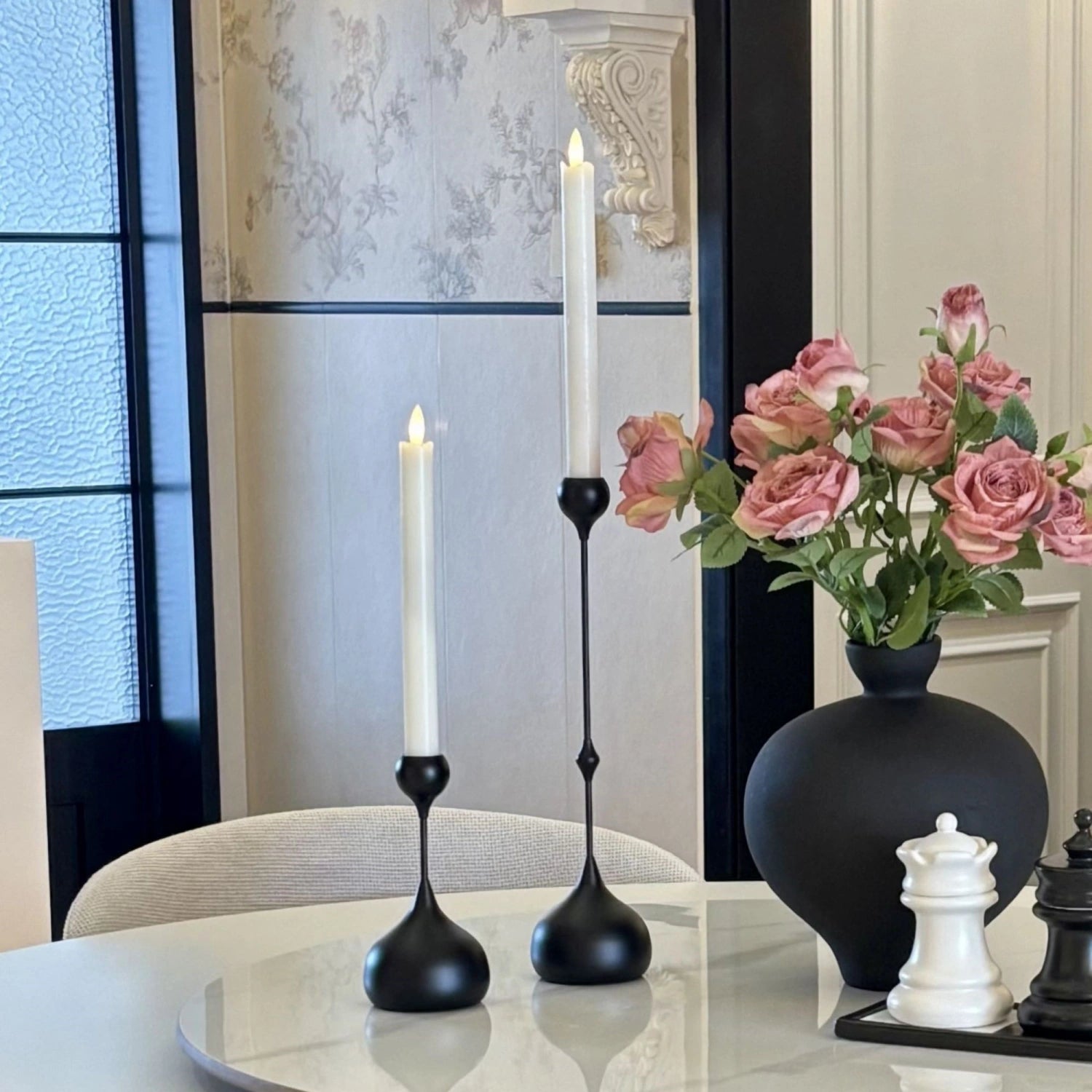 Decorative setup with black candle holders, white candles, pink flowers in a vase, and chess pieces on a table.