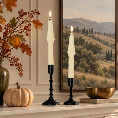 Two lit cream ghost-shaped taper candles in black metal holders, displayed on a mantel with a pumpkin, autumn leaves, and a scenic window view in the background.