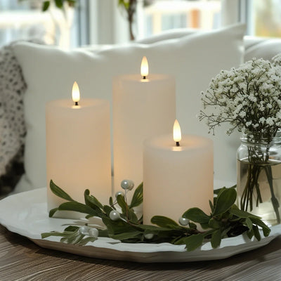 Three lit candles on a decorative tray with greenery and a vase of white flowers in the background.