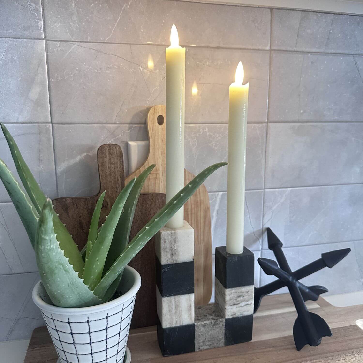 Decorative candles on a stand with a potted aloe vera plant and wooden cutting board against a tiled wall.