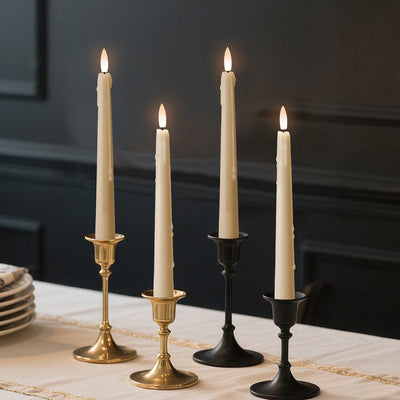 A set of three flameless taper candles in gold, black, and bronze colors, with a lifelike glow, displayed on a table.
