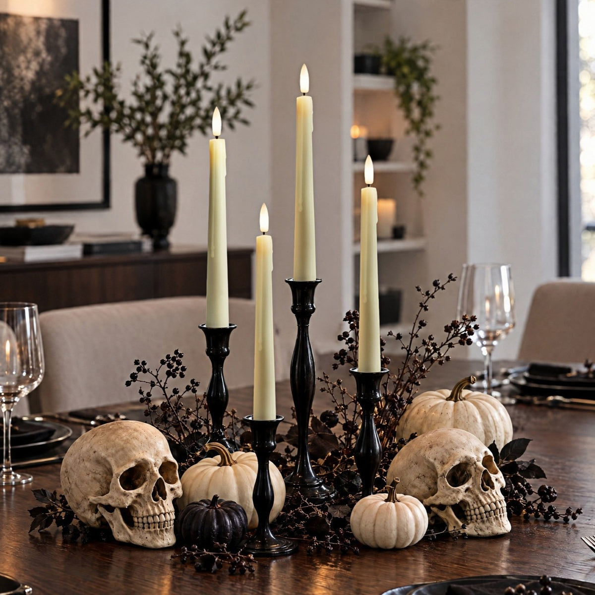 A Halloween-themed table centerpiece on a dark wood dining table, featuring two human skulls, small pumpkins, black candlesticks with cream taper candles, and dark berries, set in a dining room.
