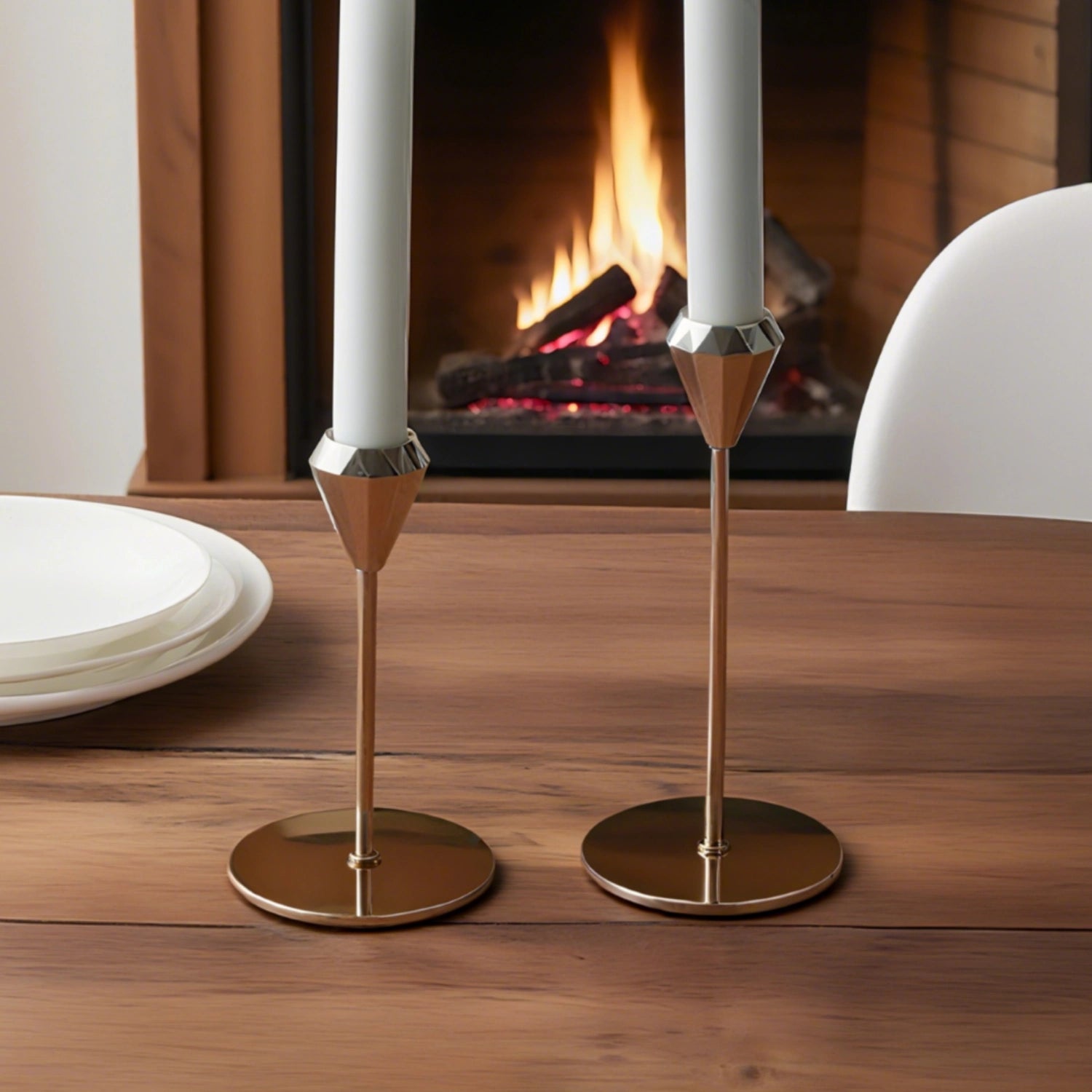 Two gold candle holders with white candles on a wooden table in front of a fireplace.