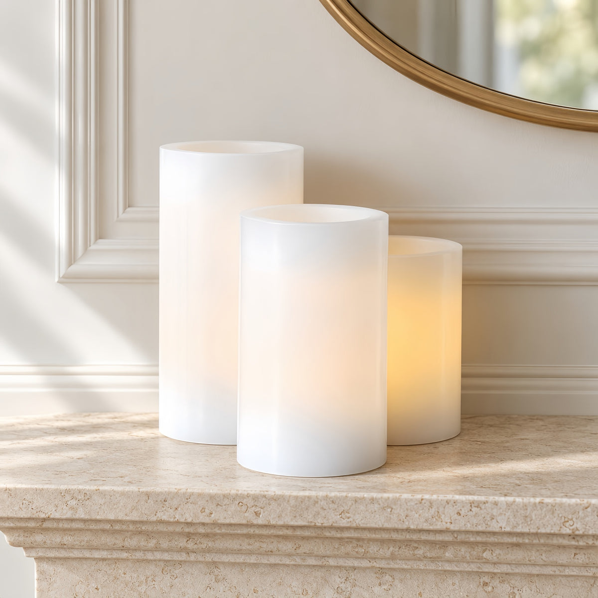 Three white pillar candles, including two unlit and one lit with a warm yellow glow, displayed on a beige stone mantelpiece in front of a cream-colored wall with molding and a gold-framed mirror.#color_white
