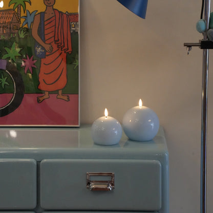 Two white candles on a light blue surface with a colorful painting in the background.