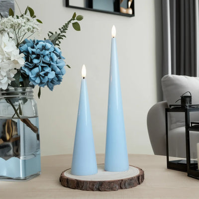 Two light blue cone-shaped candles on a wooden stand with flowers in the background.