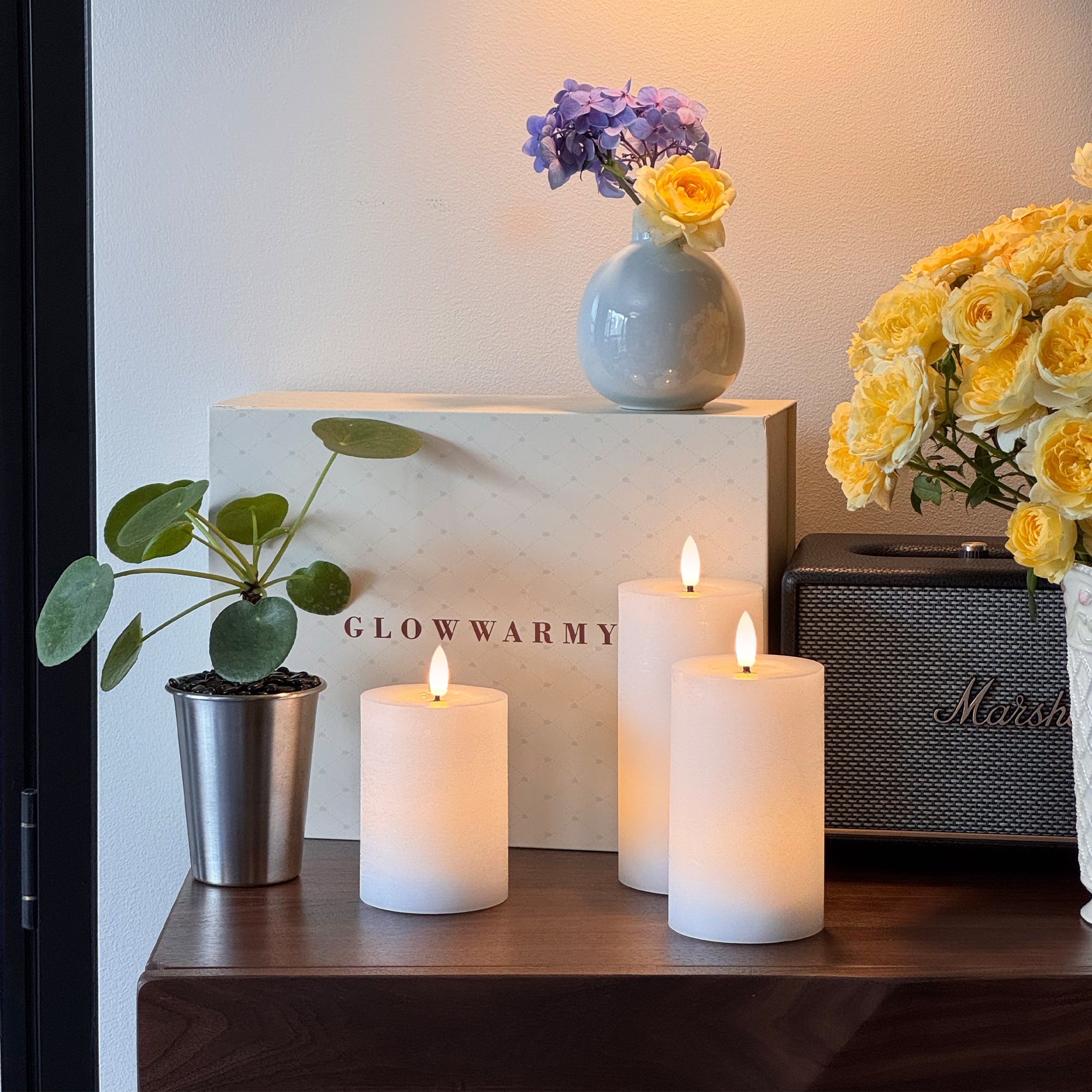 Candle holders with lit candles on a surface, featuring a 'Glow Warmly' box and decorative items.
