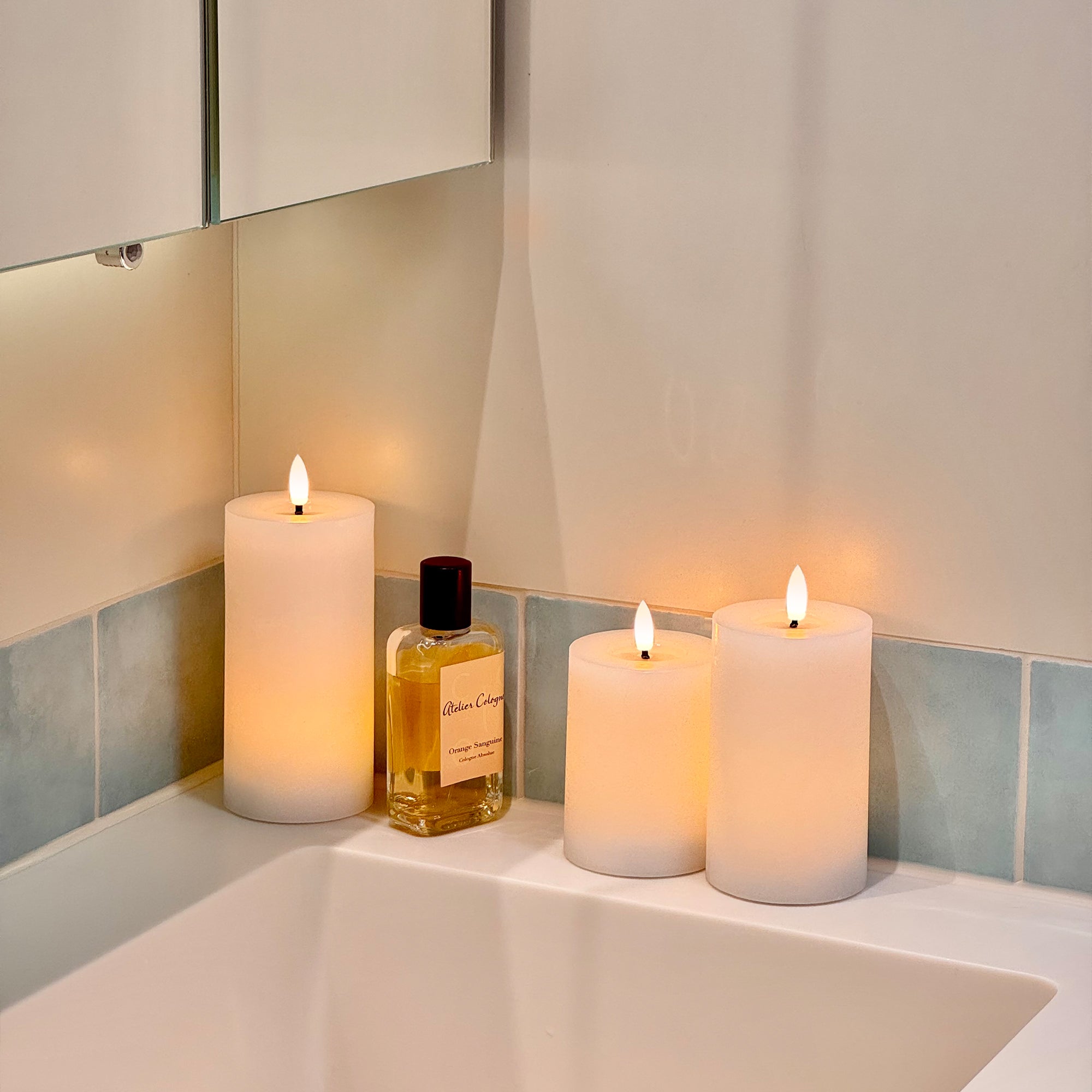 Two lit candles and a bottle of perfume on a bathroom counter with a mirror in the background.