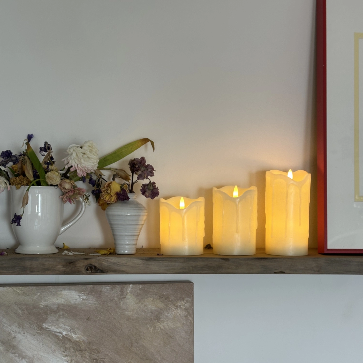 Flickering candles on a wooden shelf with flowers and a framed picture in the background.