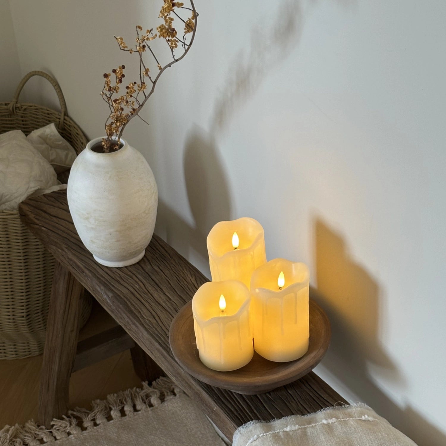 Three lit candles on a wooden surface with a vase and basket in the background.