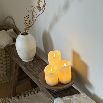 Three lit candles on a wooden surface with a vase and basket in the background.