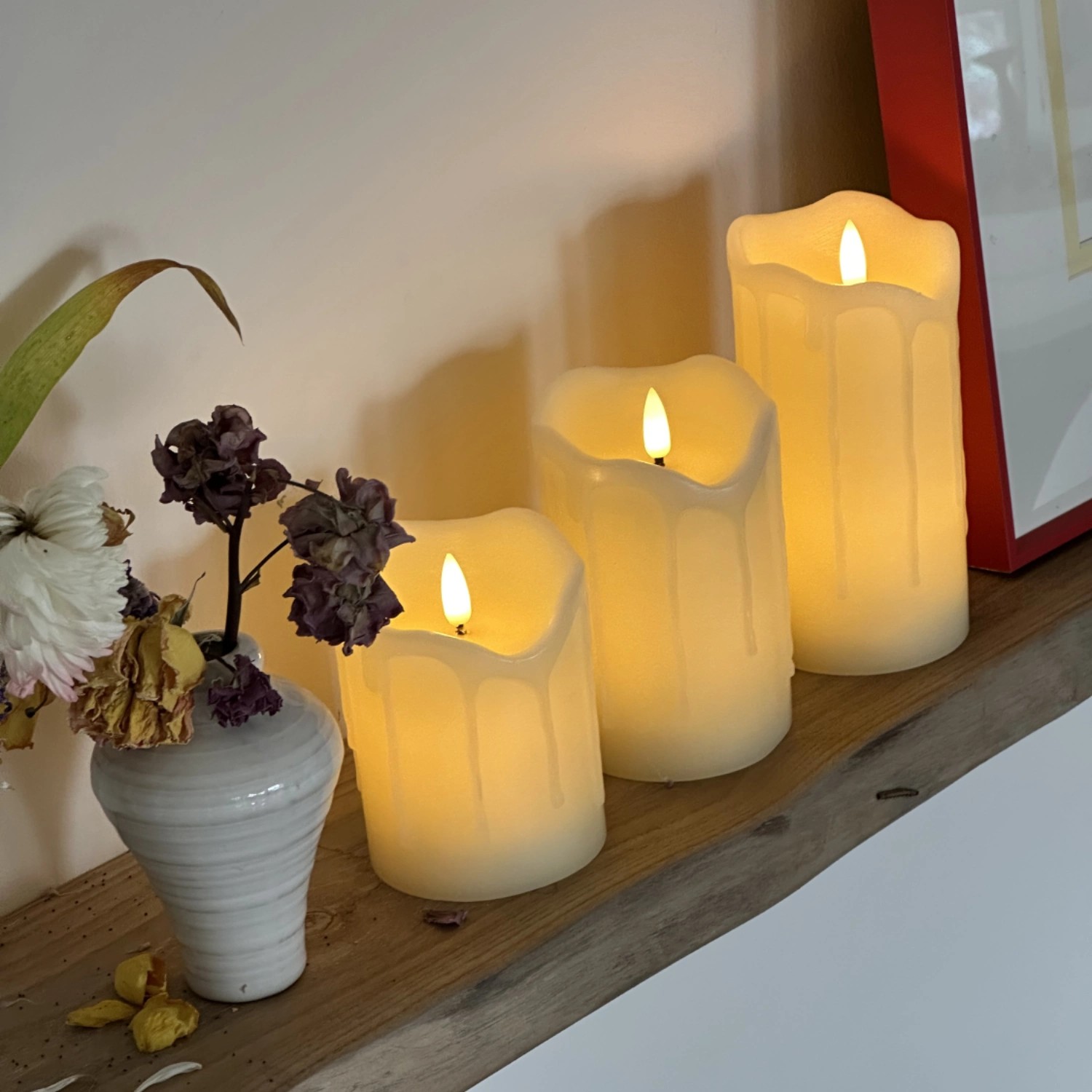 Three lit candles on a wooden shelf with a vase of flowers and a framed picture in the background.#color_ivory
