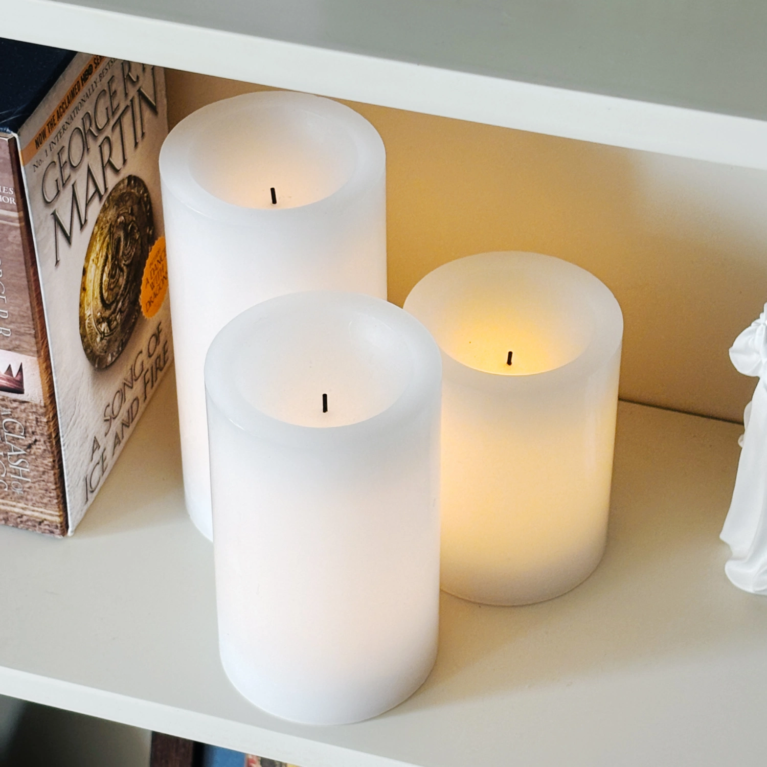 Three white candles on a shelf with a book and decorative item in the background