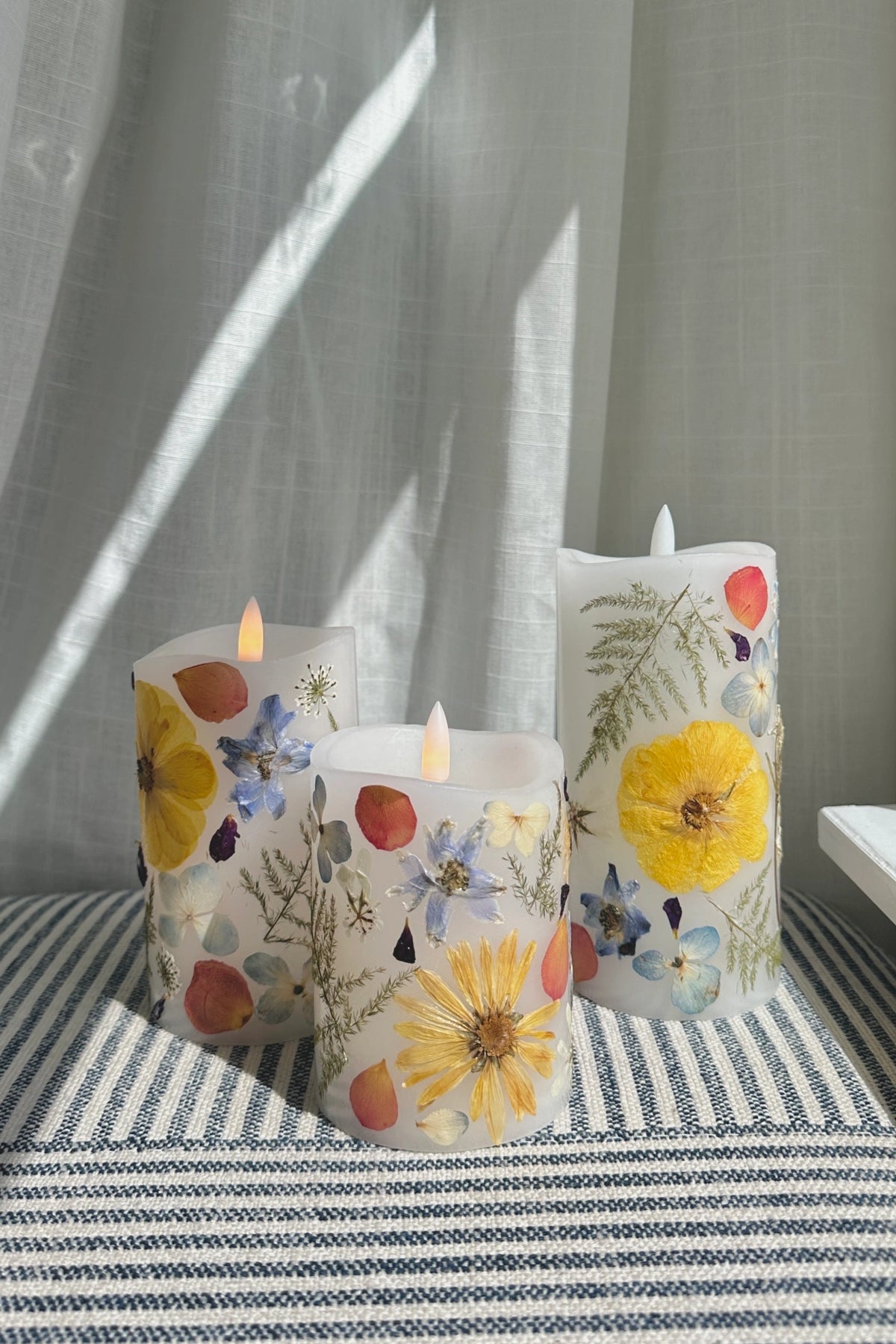 Three decorative pillar candles with a white base, decorated with colorful pressed flowers and botanical elements, arranged on a striped textile with soft window light casting shadows.