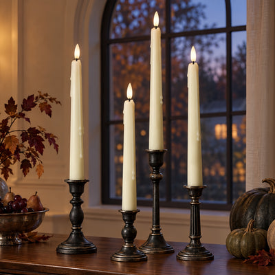 Four cream taper candles in black metal candlesticks arranged on a wooden table in a warmly lit room, with autumn leaves, a bowl of fruit, and pumpkins nearby and a window in the background.