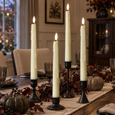 Four white taper candles in black metal candlesticks arranged on a dining table with autumn decorations, including pumpkins, berries, and a runner, in a warmly lit home interior.