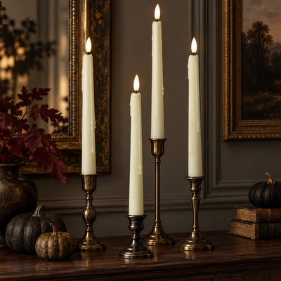 Four lit taper candles in ornate brass and silver candlesticks arranged on a wooden table in a styled interior setting with pumpkins, framed art, and books.